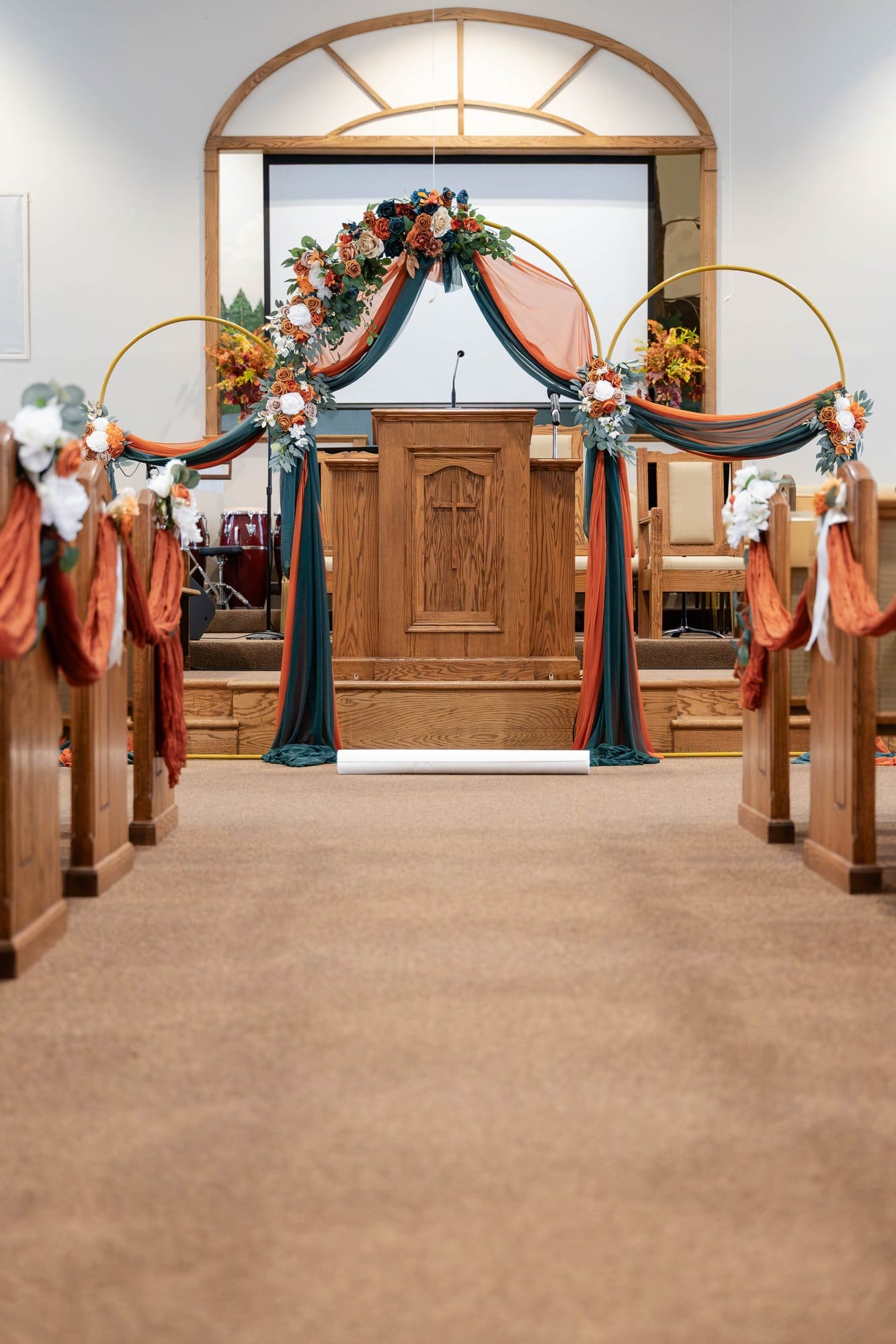 View down the church aisle toward the gold hoop arch, with pews swagged in copper gauze and white roses