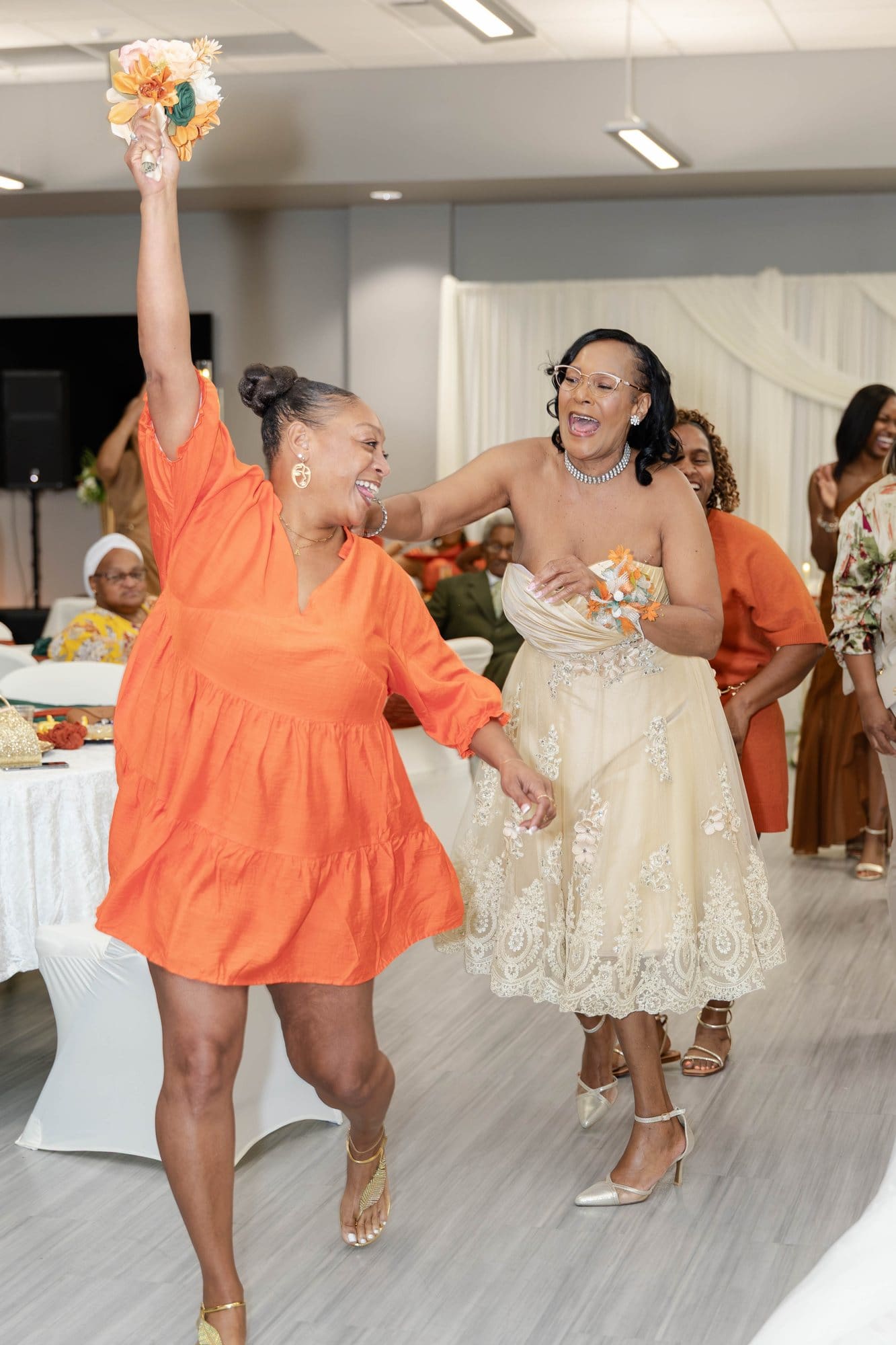 A guest in an orange dress raising the caught bouquet as another guest laughs beside her