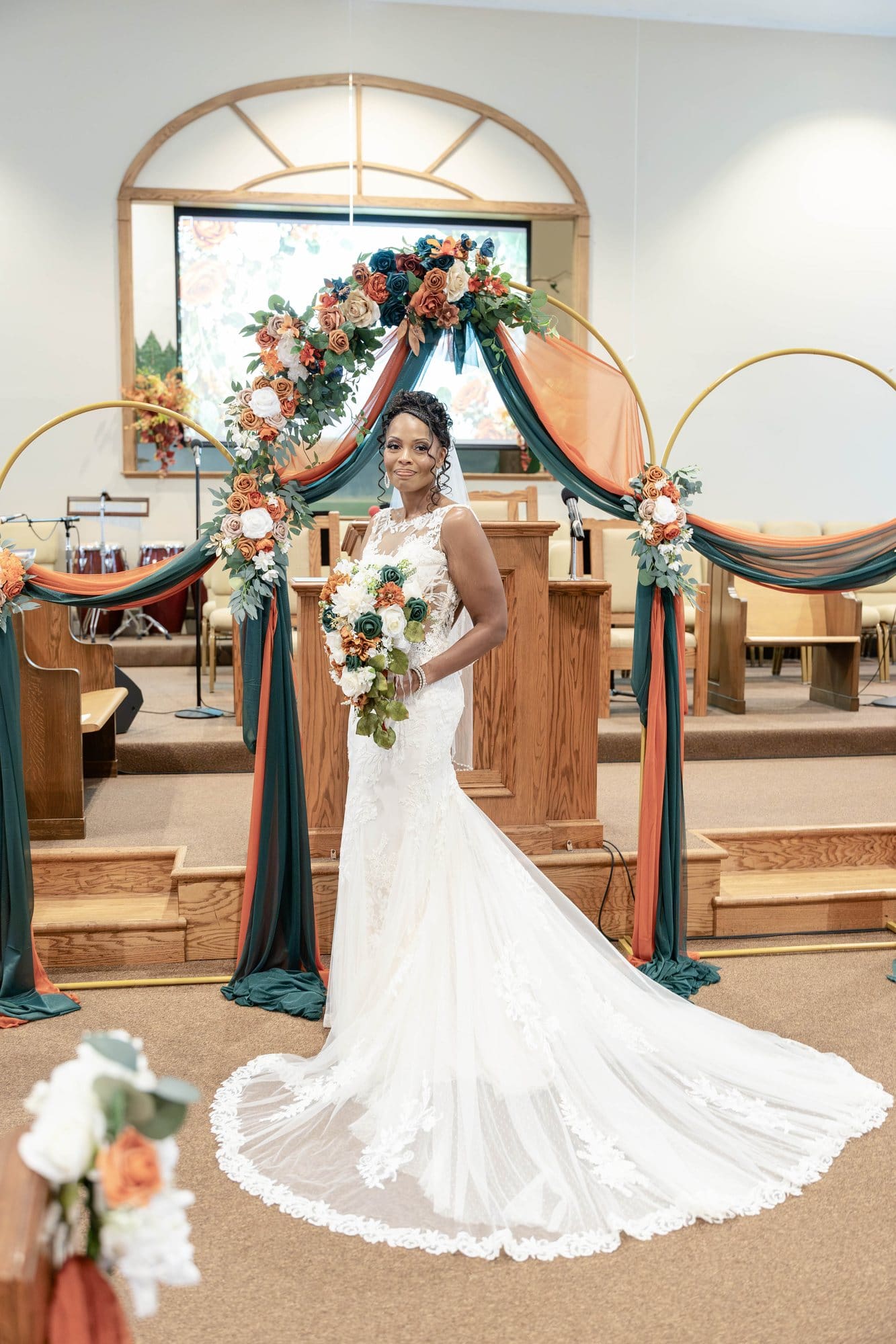 Nikeesha standing at the altar in her ivory lace gown with a long tulle train, holding her emerald and copper bouquet