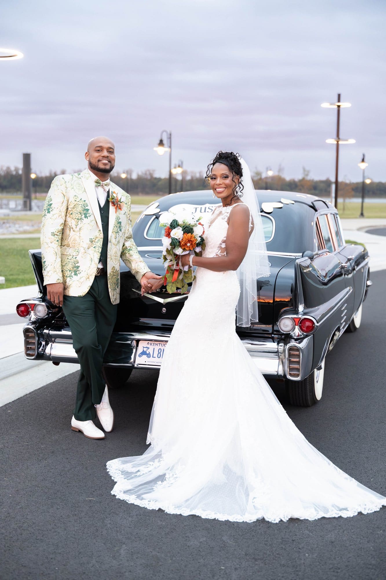 David and Nikeesha leaning against the back of a vintage Cadillac at dusk under a purple and pink sky