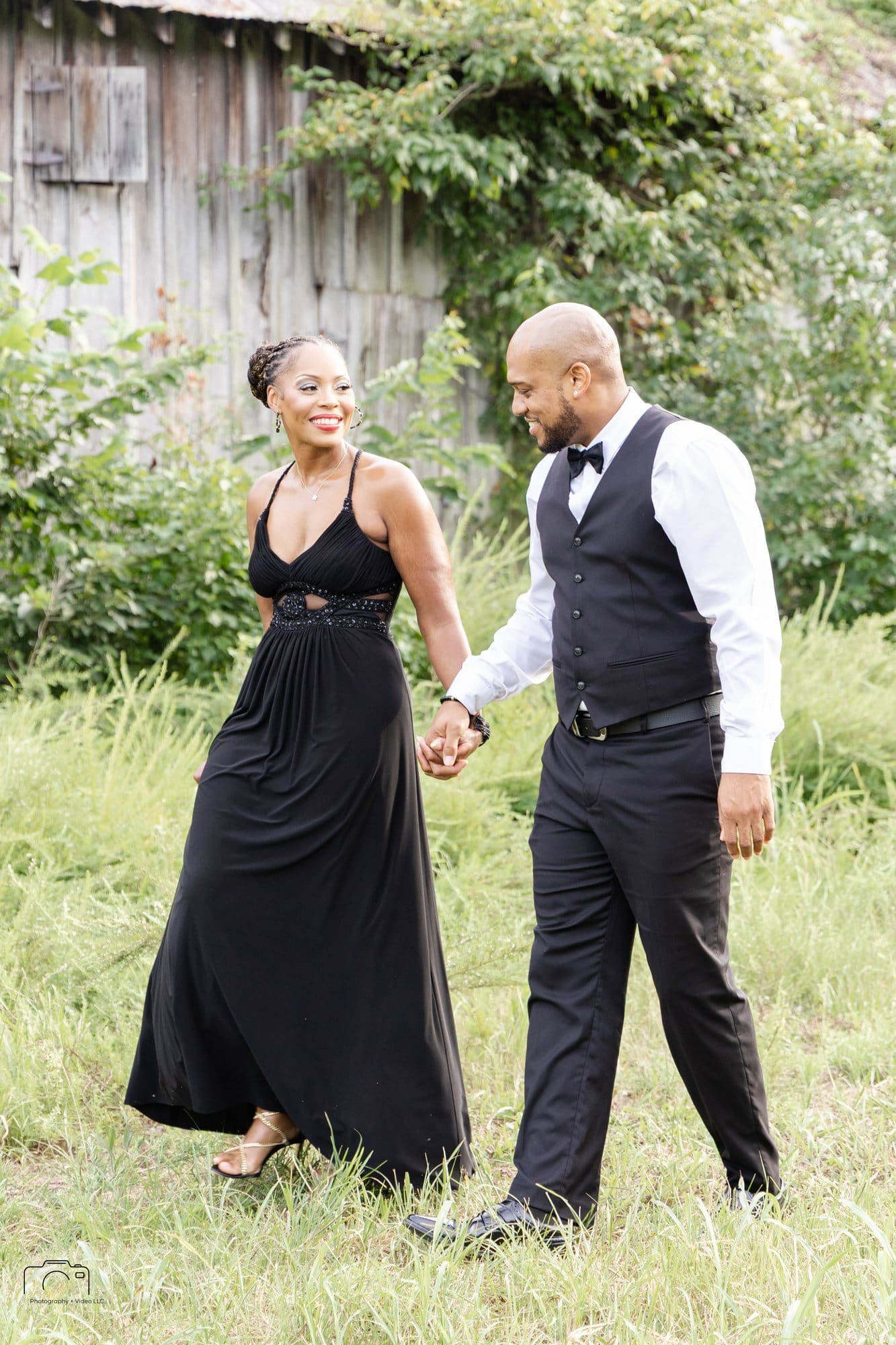 David and Nikeesha in formal black attire walking hand in hand through a grassy field beside an old barn at Empire Point, Land Between the Lakes