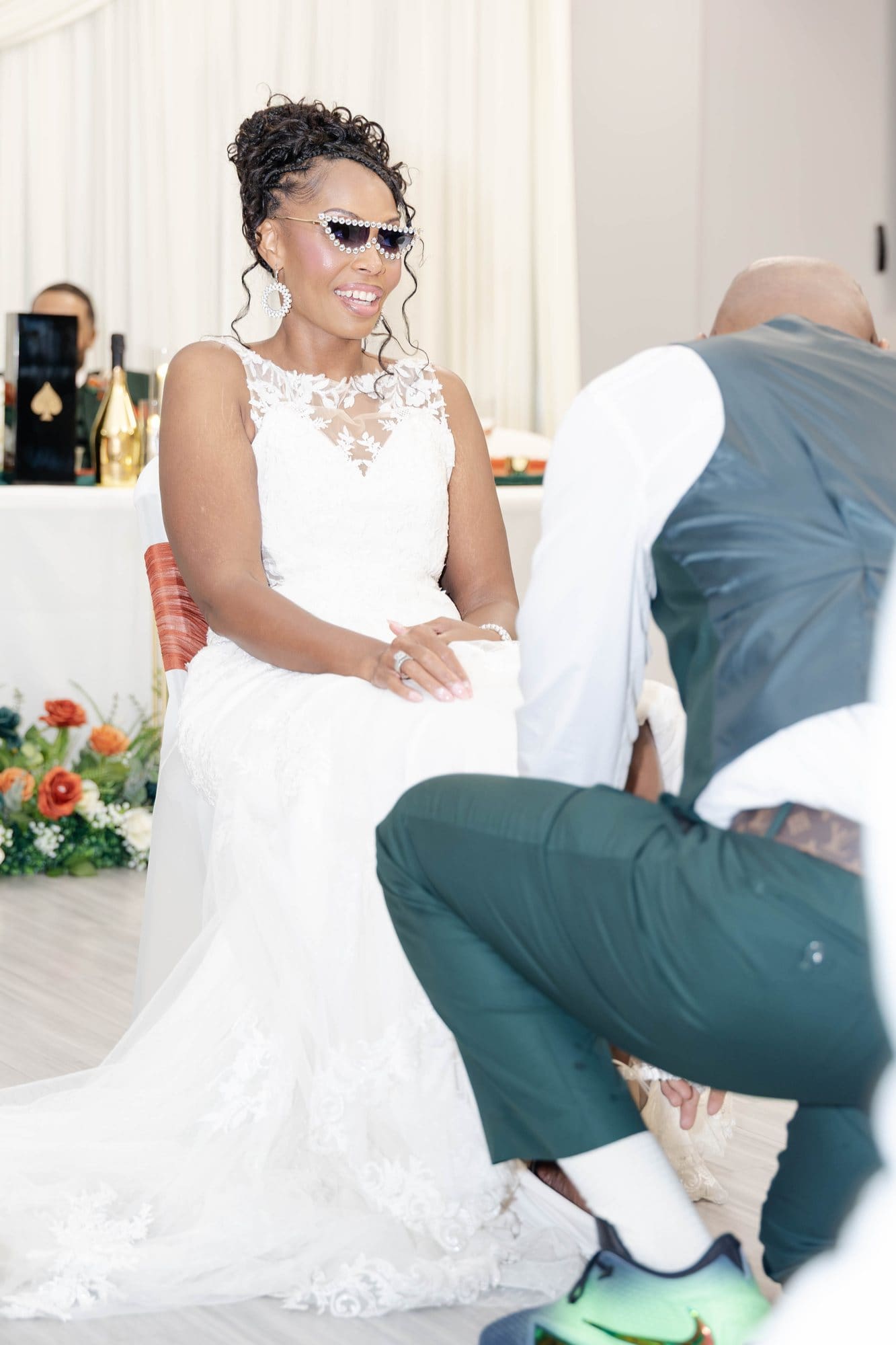 Nikeesha seated in sunglasses while David kneels for the garter toss
