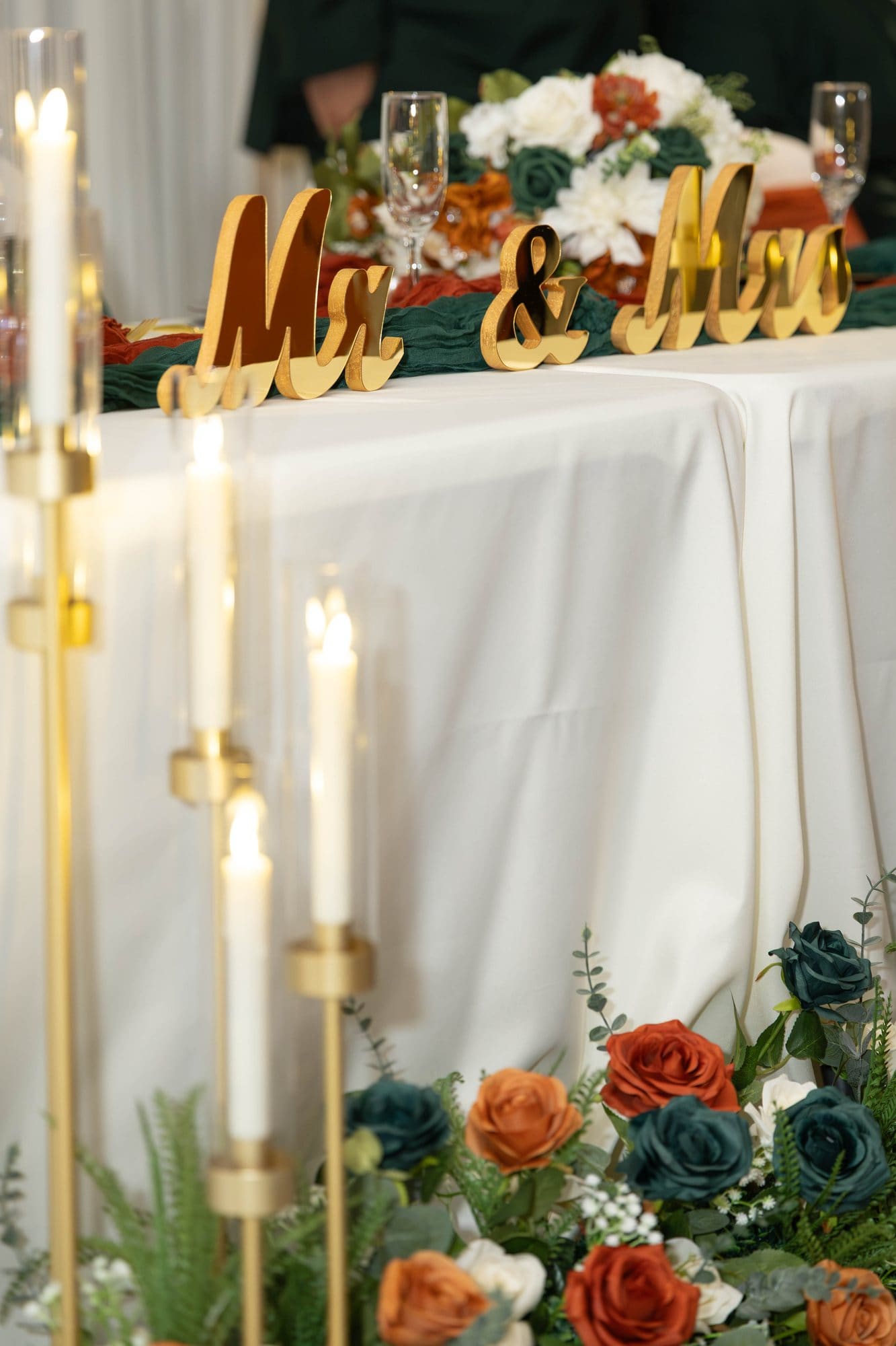 Gold Mr. & Mrs. sign beside tall candles and roses in copper, cream and emerald