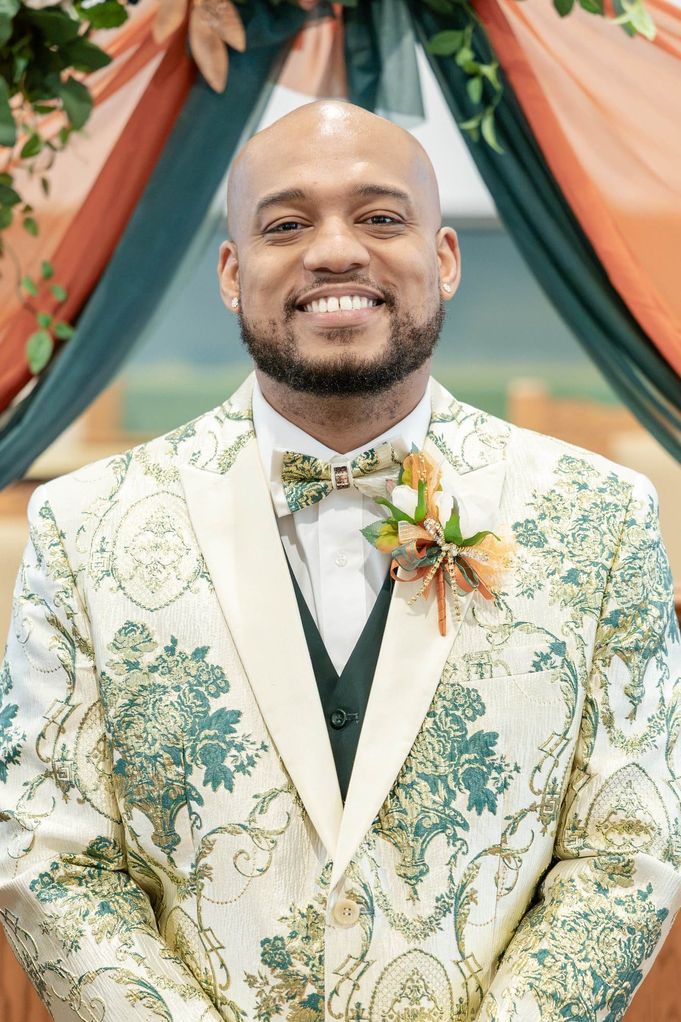 David smiling in his ivory brocade tuxedo jacket woven with emerald and gold beneath the ceremony arch