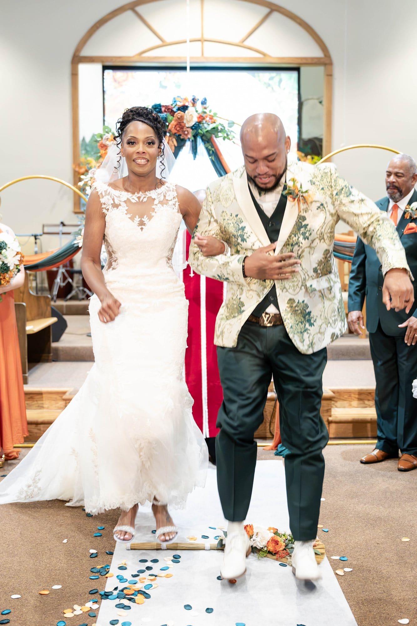 David and Nikeesha jumping the broom together in the church aisle strewn with petals