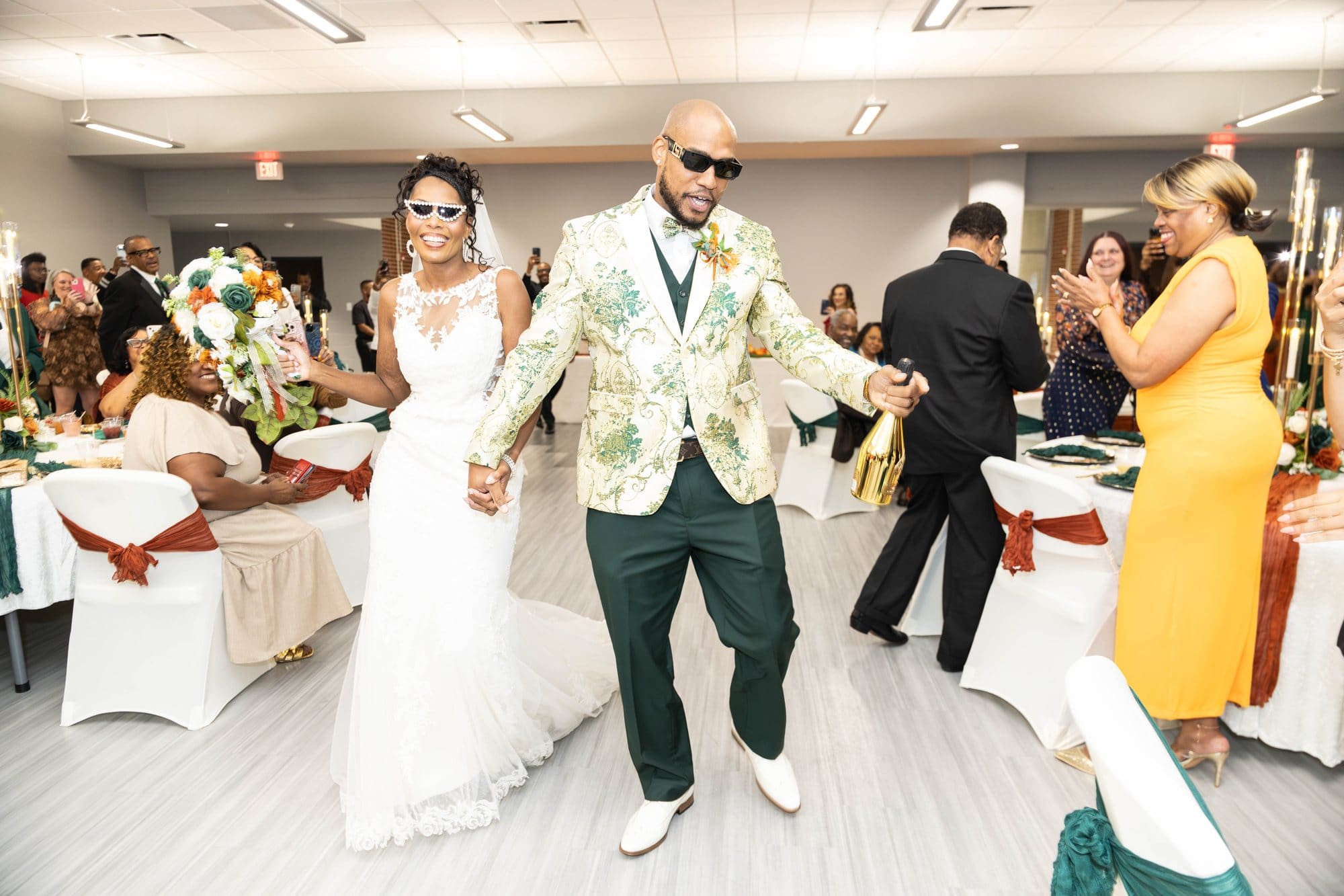 David and Nikeesha entering their reception in sunglasses, hand in hand, with her bouquet raised high and David holding a gold bottle