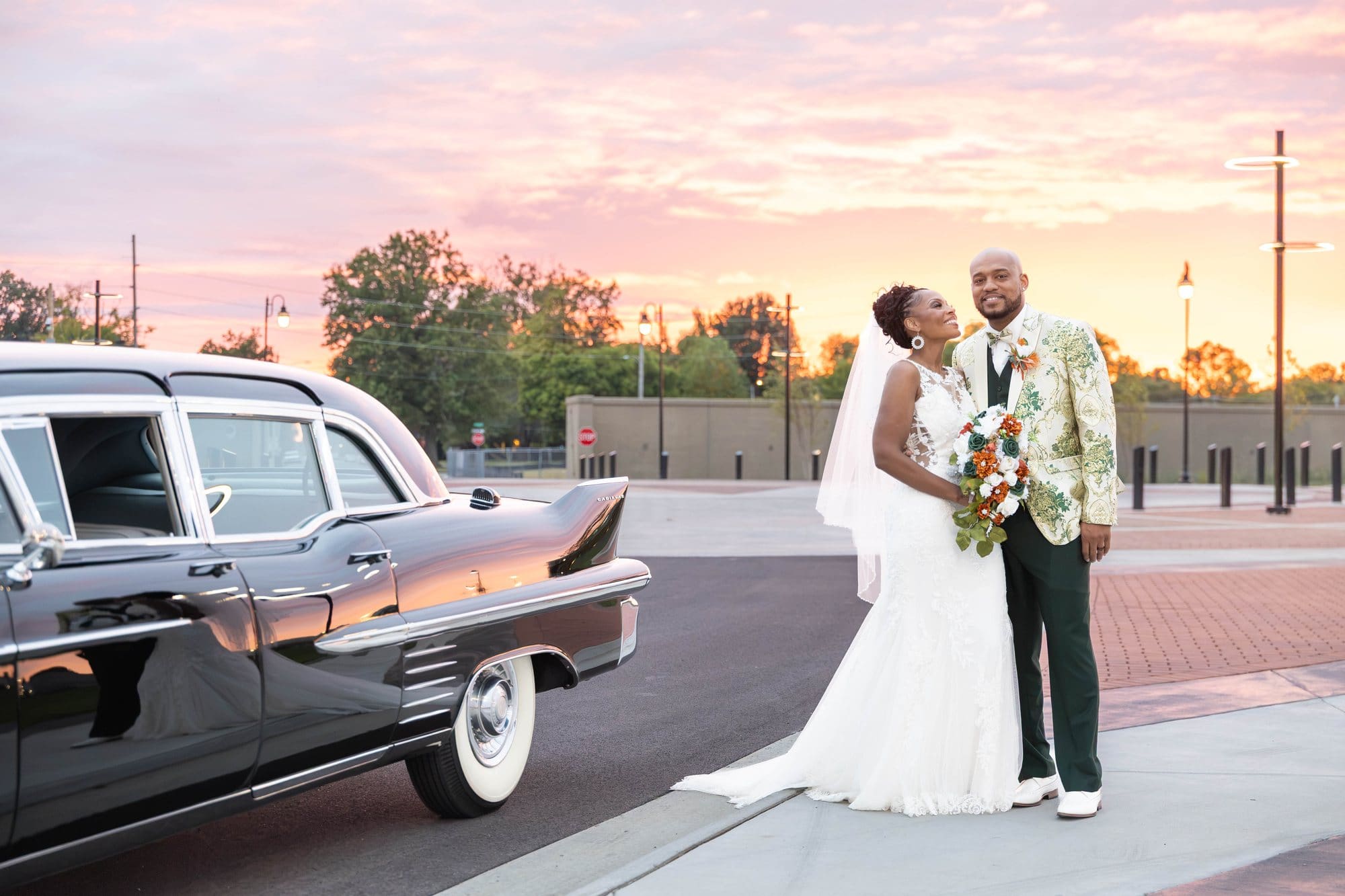 Nikeesha and David smiling at each other beside a vintage Cadillac under a pink and orange sunset sky in downtown Paducah, KY