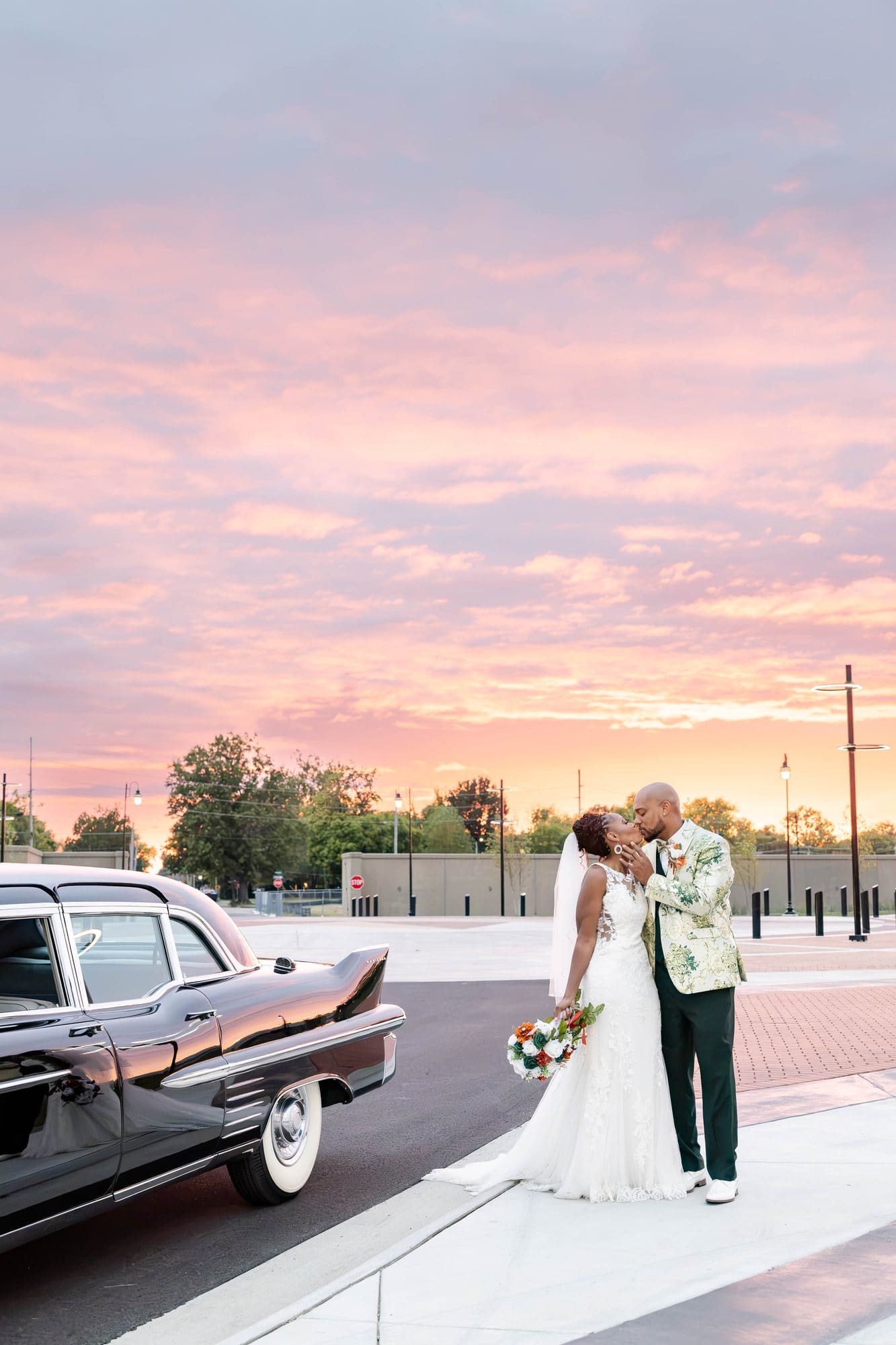 Wide view of David and Nikeesha kissing beside the Cadillac below a sky streaked with pink and orange