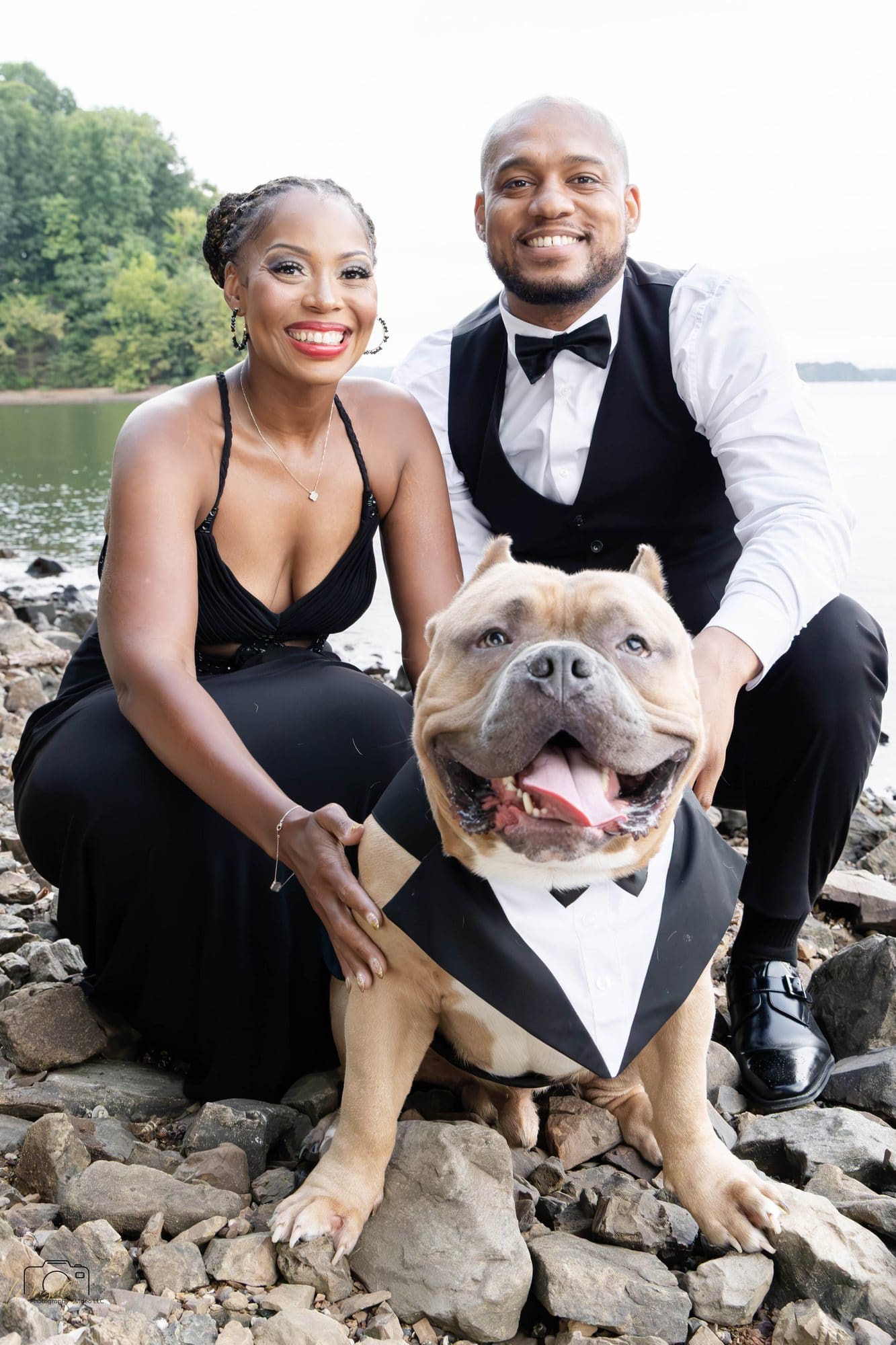 David and Nikeesha crouching on a rocky shore with their dog Veno in a tuxedo at Kentucky Lake